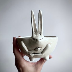 May include: White ceramic bowl shaped like a rabbit with its arms crossed and long ears. The bowl has a glazed finish and is hand-painted with black details.
