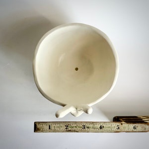 May include: White ceramic planter shaped like two hands holding a bowl. The bowl has a drainage hole in the bottom.