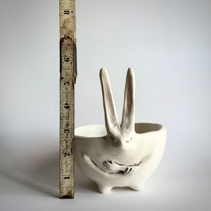 May include: White ceramic planter shaped like a rabbit with its arms crossed in front of it. The planter has a wide, shallow bowl.