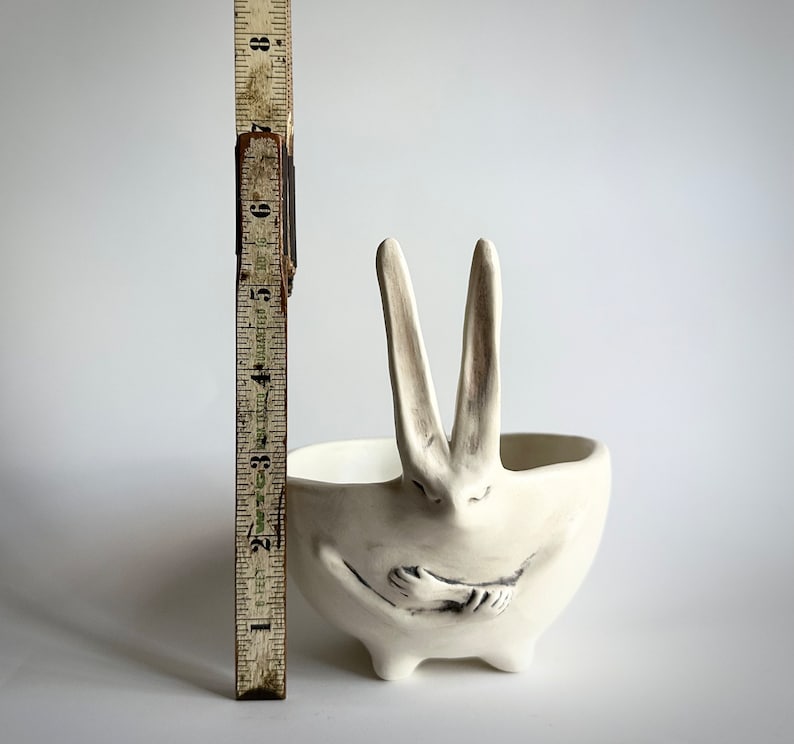 May include: White ceramic planter shaped like a rabbit with its arms crossed in front of it. The planter has a wide, shallow bowl.