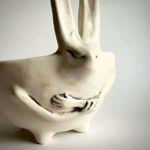 May include: White ceramic planter shaped like a rabbit holding its arms around itself. The planter has a bowl-shaped top and three legs.