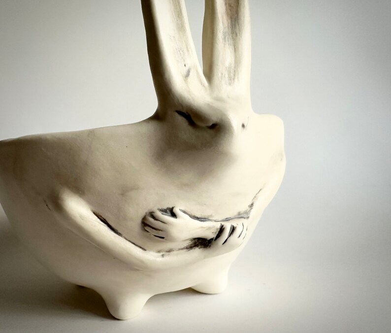 May include: White ceramic planter shaped like a rabbit holding its arms around itself. The planter has a bowl-shaped top and three legs.
