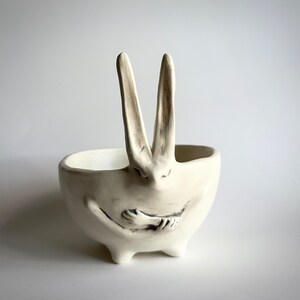 May include: White ceramic bowl shaped like a rabbit with its arms crossed in front of it. The rabbit has long ears and a small, round nose.