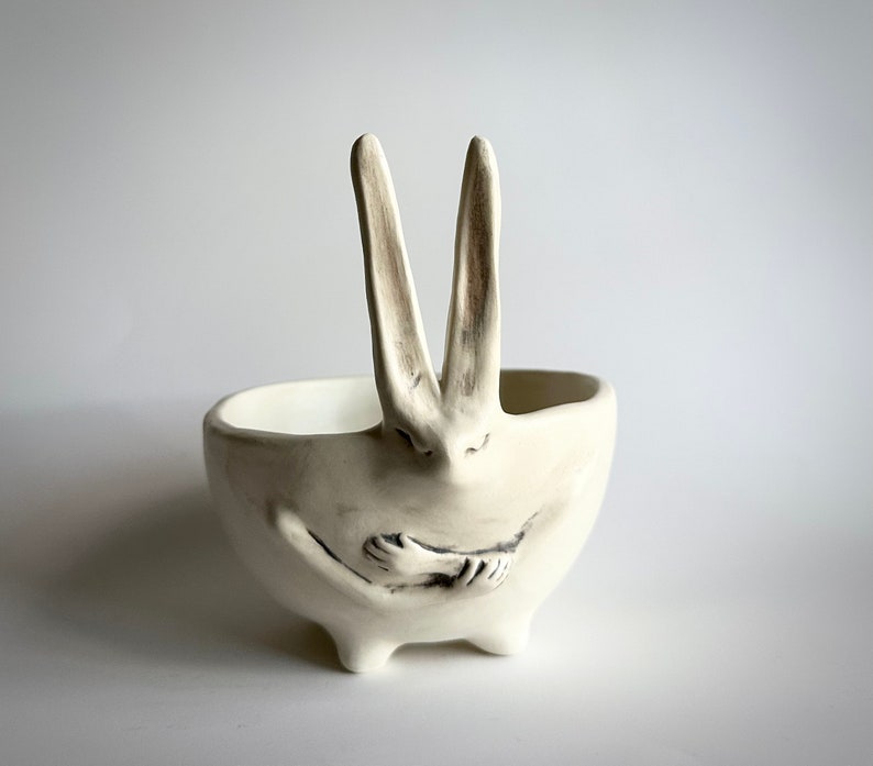 May include: White ceramic bowl shaped like a rabbit with its arms crossed in front of it. The rabbit has long ears and a small, round nose.