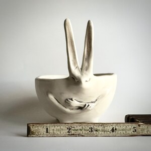 May include: White ceramic bowl shaped like a rabbit with its arms crossed in front of it. The rabbit's ears are upright and the bowl is sitting on a wooden ruler.