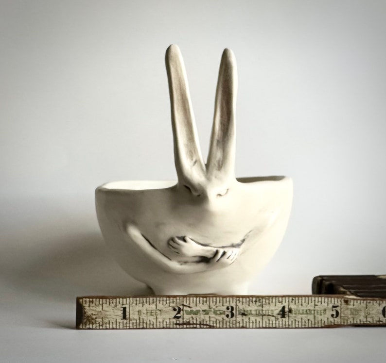 May include: White ceramic bowl shaped like a rabbit with its arms crossed in front of it. The rabbit's ears are upright and the bowl is sitting on a wooden ruler.