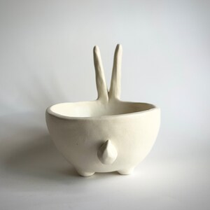 May include: White ceramic bowl shaped like a rabbit with two long ears and a small tail. The bowl is sitting on a white surface.