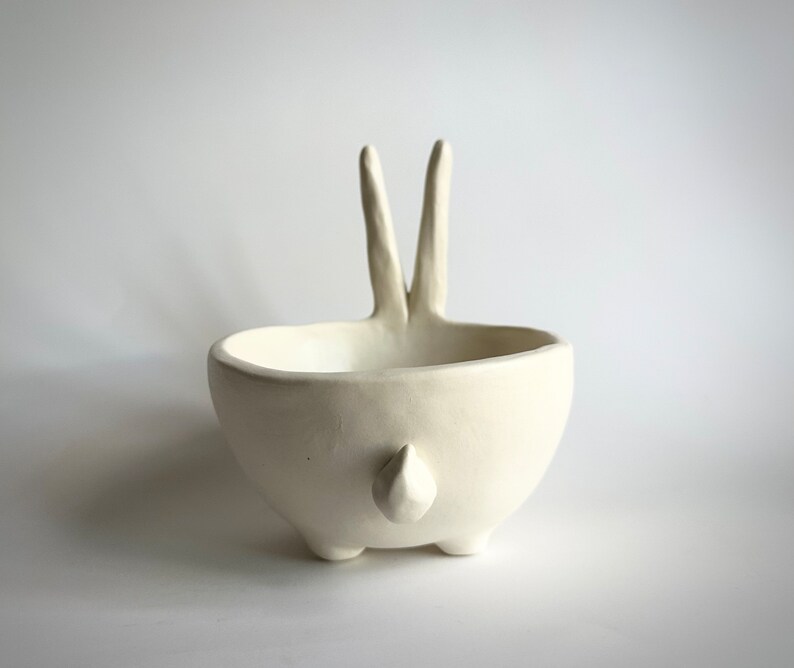 May include: White ceramic bowl shaped like a rabbit with two long ears and a small tail. The bowl is sitting on a white surface.