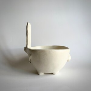 May include: White ceramic bowl shaped like a rabbit with a long ear standing upright. The bowl has four legs and two small arms.