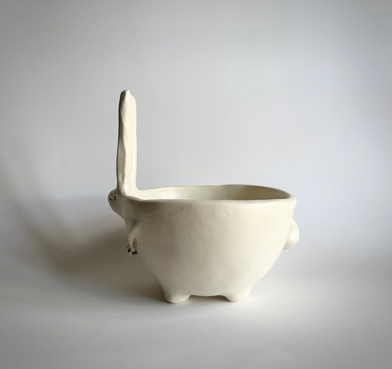 May include: White ceramic bowl shaped like a rabbit with a long ear standing upright. The bowl has four legs and two small arms.