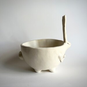 May include: White ceramic bowl shaped like a rabbit with its ears standing up. The bowl has four legs and a rounded body.