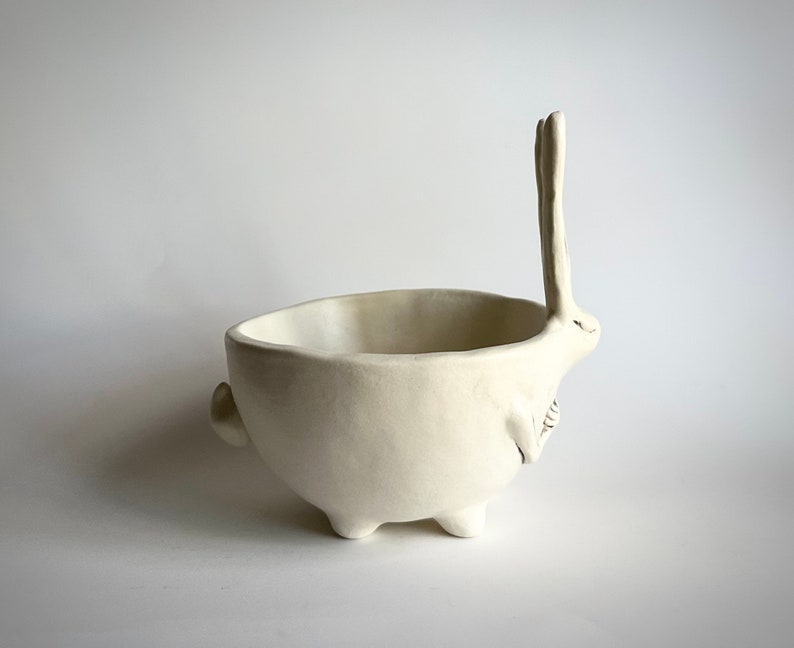 May include: White ceramic bowl shaped like a rabbit with its ears standing up. The bowl has four legs and a rounded body.