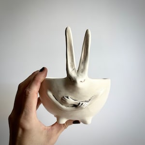 May include: White ceramic planter shaped like a rabbit with its arms crossed in front of its chest. The planter has two long ears and a small, round base.