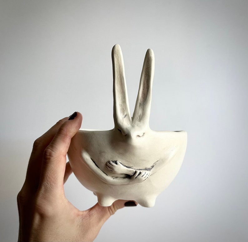 May include: White ceramic planter shaped like a rabbit with its arms crossed in front of its chest. The planter has two long ears and a small, round base.