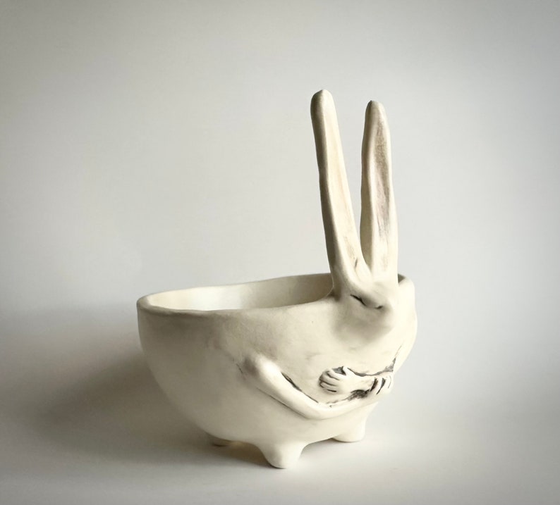 May include: White ceramic planter shaped like a rabbit with its arms crossed in front of it. The planter has a bowl-shaped top and four small feet.