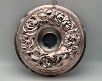 Puede incluir: Un molde para bizcocho bundt de color cobre con un diseño de rosas. El molde tiene forma circular con un agujero central y un patrón floral decorativo en los bordes. Incluye un pequeño lazo para colgar.