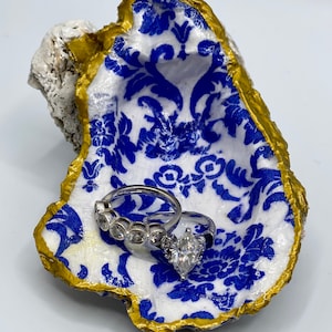 May include: A decorative oyster shell ring dish. The interior features a blue and white floral pattern, with a gold painted rim. Two silver rings with clear stones rest inside the dish.