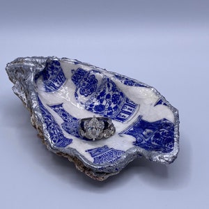 Op de afbeelding: Een decoratieve oesterschaal met een blauw en wit porseleinen patroon. De schelp heeft een zilverkleurige rand en bevat een zilveren ring met een grote, heldere edelsteen en kleinere stenen.