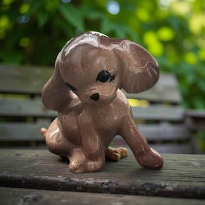 May include: A brown ceramic puppy figurine with large ears and black accents. The puppy is in a seated position, looking down. The figurine is on a wooden surface with a blurred green background.