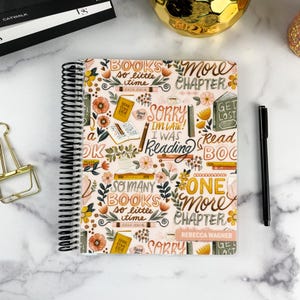 May include: A spiral-bound notebook with a cream-colored cover featuring a repeating pattern of books, flowers, and text including "Books so little time," "Sorry I'm late, I was reading," "One more chapter," and the name "Rebecca Wagner" printed at the bottom.