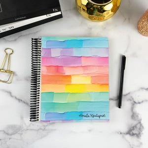 May include: A spiral-bound notebook featuring a cover with horizontal, textured brushstrokes in a rainbow gradient of blue, purple, pink, orange, yellow, and teal. The name Amala Koutapuri is printed in black script in the bottom right corner.