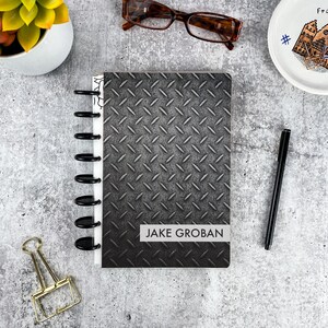 May include: A disc-bound notebook featuring a dark gray diamond plate metal pattern on the cover. A white rectangular label on the lower portion of the cover displays the name JAKE GROBAN in black capital letters. The notebook has black plastic discs.