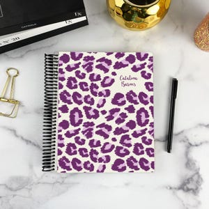 May include: A spiral-bound notebook featuring a purple and light purple leopard print pattern on a white background. The cover includes the printed name Catalina Barnes in a dark purple script font. A black metal spiral coil binds the pages.