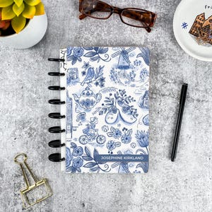 May include: A disc-bound notebook featuring a blue and white pattern of Dutch-inspired illustrations including windmills, clogs, a teapot, and a person on a bicycle. The cover includes the name Josephine Kirkland printed on a blue rectangular label.