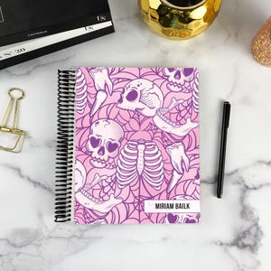 May include: A spiral-bound notebook featuring a repeating pattern of skulls, rib cages, and teeth in shades of pink and purple. The cover includes a white rectangular label with the black printed text MIRIAM BAILK on the bottom right corner.