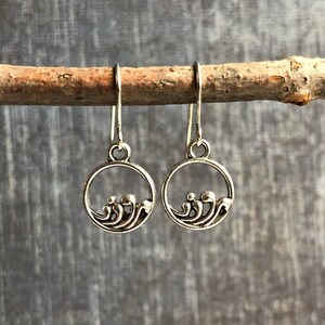 May include: Silver earrings with a circle design featuring a wave pattern. The earrings are hanging from a branch.
