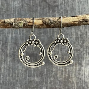 May include: Silver tone earrings with a floral design. The earrings are circular and have a delicate, intricate design.