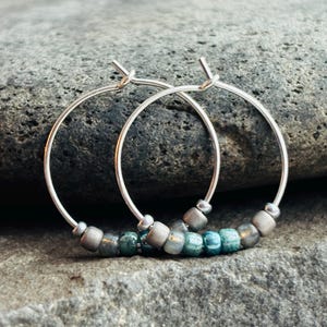 May include: A pair of silver hoop earrings with a row of small, gray and teal beads. The earrings are displayed on a gray stone surface, with a blurred wooden background.
