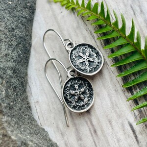 May include: A pair of silver earrings with a textured, circular design. Each earring features a detailed, raised floral pattern in the center. The earrings hang from simple, curved ear wires, showcasing a rustic, nature-inspired aesthetic.