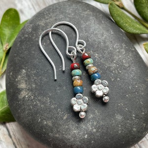 May include: A pair of dangle earrings with silver hooks. Each earring features a strand of colorful beads in red, turquoise, yellow, and blue, topped with a silver flower-shaped charm. The earrings are displayed on a dark gray stone.