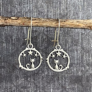 May include: Silver dangle earrings featuring a cat silhouette inside a circle with stars. The cat is looking up at the stars.