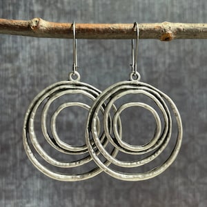 May include: A pair of silver-toned earrings with a layered, circular design. Each earring features three concentric circles, creating a textured, openwork effect. The earrings hang from simple, curved ear wires, showcasing a rustic, bohemian style.