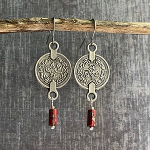 May include: Silver earrings with a round coin-shaped charm featuring Arabic script and a red stone dangle.