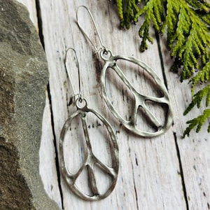 May include: A pair of silver-toned earrings featuring a peace sign design within an oval frame. The earrings have a long hook for pierced ears. The background includes a wooden surface and green foliage.