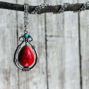 Peut inclure: Un collier en cha&icirc;ne argent&eacute;e avec un pendentif en pierre rouge et blanche. Le pendentif a la forme d'une larme et est dot&eacute; d'un cadre argent&eacute; avec des d&eacute;tails d&eacute;coratifs. Le collier est suspendu &agrave; une branche.