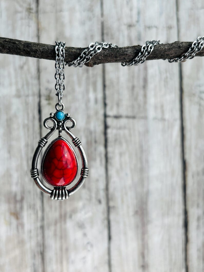 Peut inclure: Un collier en cha&icirc;ne argent&eacute;e avec un pendentif en pierre rouge et blanche. Le pendentif a la forme d'une larme et est dot&eacute; d'un cadre argent&eacute; avec des d&eacute;tails d&eacute;coratifs. Le collier est suspendu &agrave; une branche.