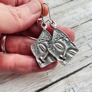 May include: A pair of silver earrings with a textured, abstract design. The earrings are shaped like a square with rounded corners and have a dangling hook.