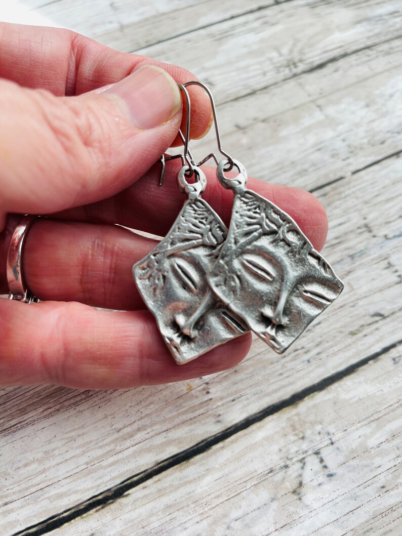 May include: A pair of silver earrings with a textured, abstract design. The earrings are shaped like a square with rounded corners and have a dangling hook.