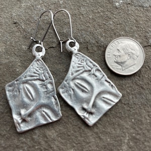 May include: A pair of silver earrings with a face design. The earrings are made of metal and have a textured surface. The earrings are about the size of a dime.