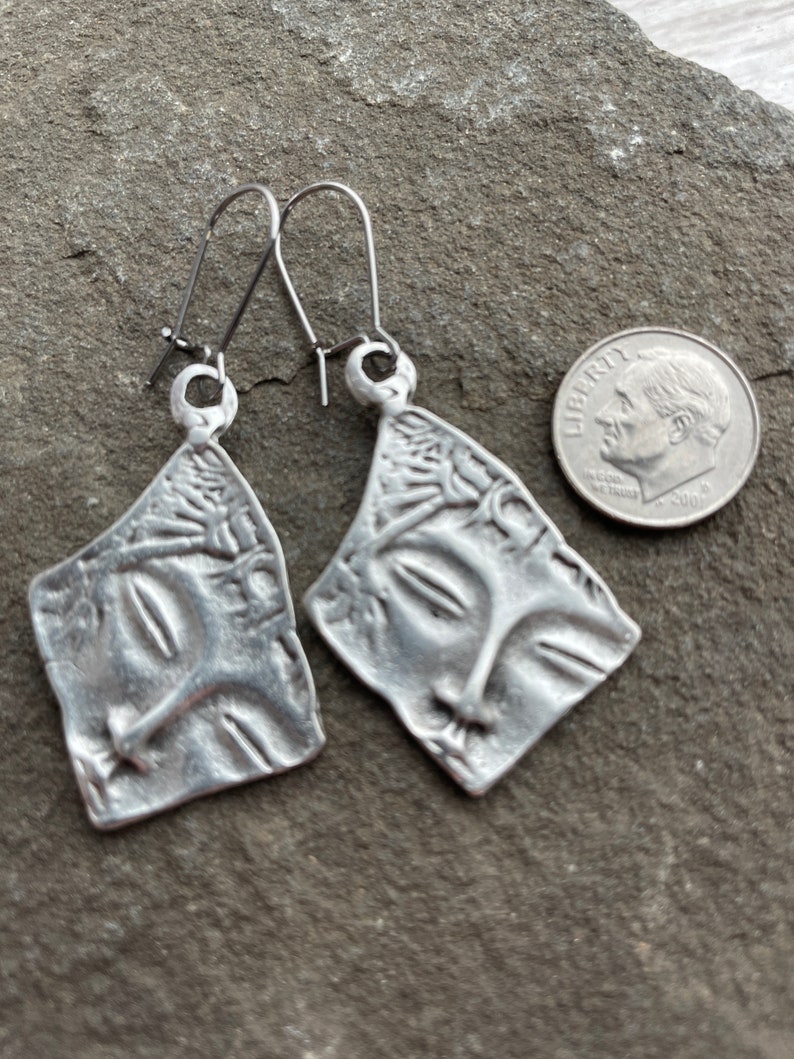 May include: A pair of silver earrings with a face design. The earrings are made of metal and have a textured surface. The earrings are about the size of a dime.