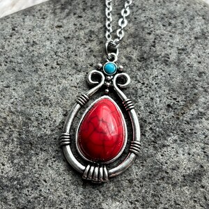 Peut inclure: Un collier en argent avec un pendentif en pierre rouge et blanche. Le pendentif est entour&eacute; d'un cadre en argent avec des d&eacute;tails complexes. Le collier est sur un fond de pierre grise.