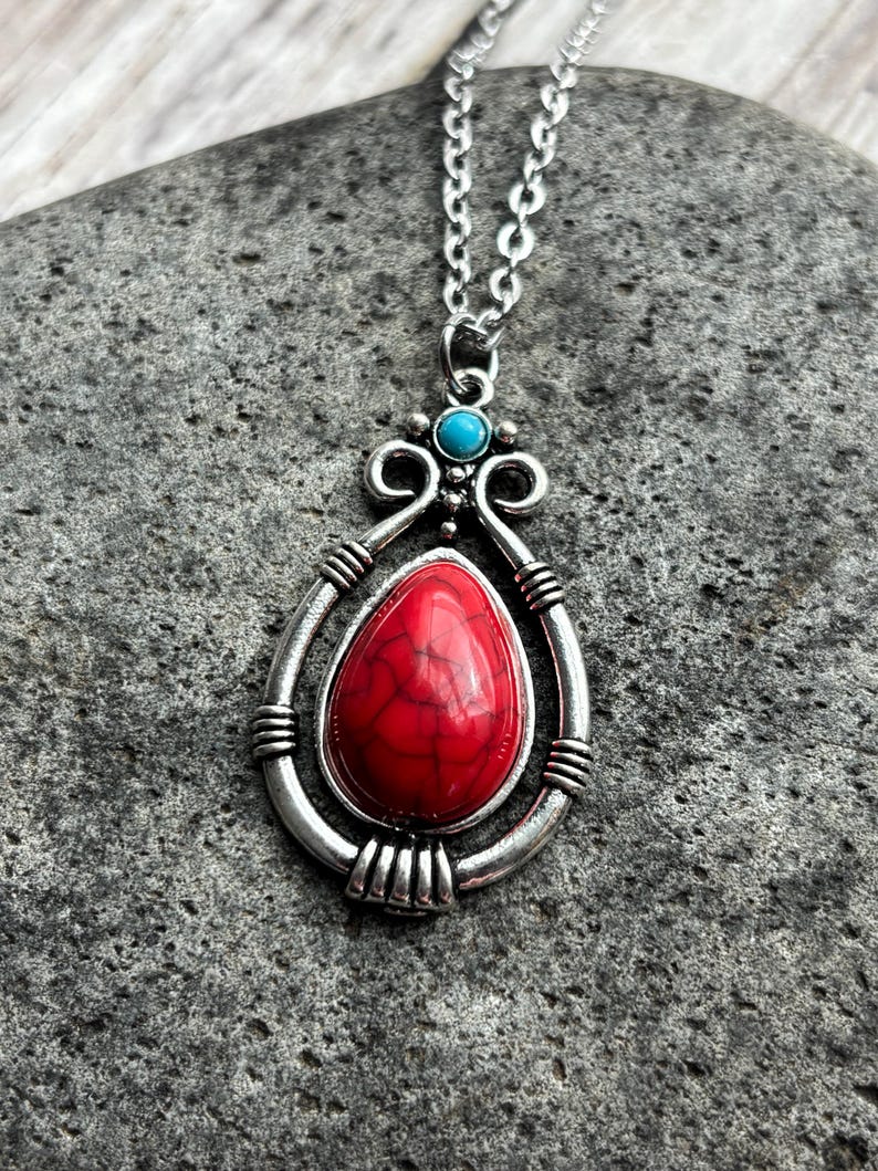 Peut inclure: Un collier en argent avec un pendentif en pierre rouge et blanche. Le pendentif est entour&eacute; d'un cadre en argent avec des d&eacute;tails complexes. Le collier est sur un fond de pierre grise.