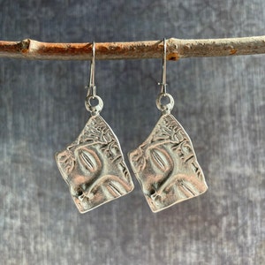 May include: Silver earrings with a textured, abstract design. The earrings are shaped like a teardrop and have a silver hook.