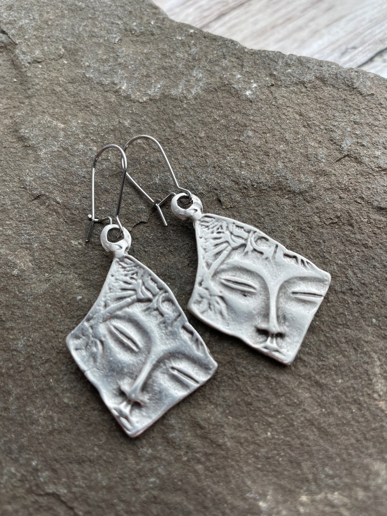 May include: A pair of silver earrings with a face design. The earrings are made of metal and have a simple, elegant design.