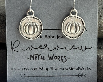 May include: A pair of silver-toned drop earrings featuring a circular charm with a raised pumpkin design. The earrings are displayed on a grey card printed with the text Riverview METAL WORKS and the website www.etsy.com/shop/RiverviewMetalWorks.