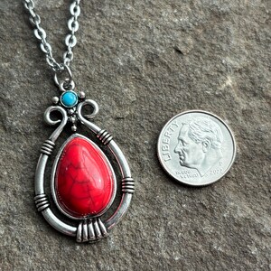 Peut inclure: Un collier en cha&icirc;ne argent avec un pendentif en pierre rouge et blanche. Le pendentif est entour&eacute; d'un cadre argent&eacute; avec une pierre turquoise en haut. Le collier est pr&eacute;sent&eacute; &agrave; c&ocirc;t&eacute; d'une pi&egrave;ce de dix centimes d'euro pour comparaison de taille.